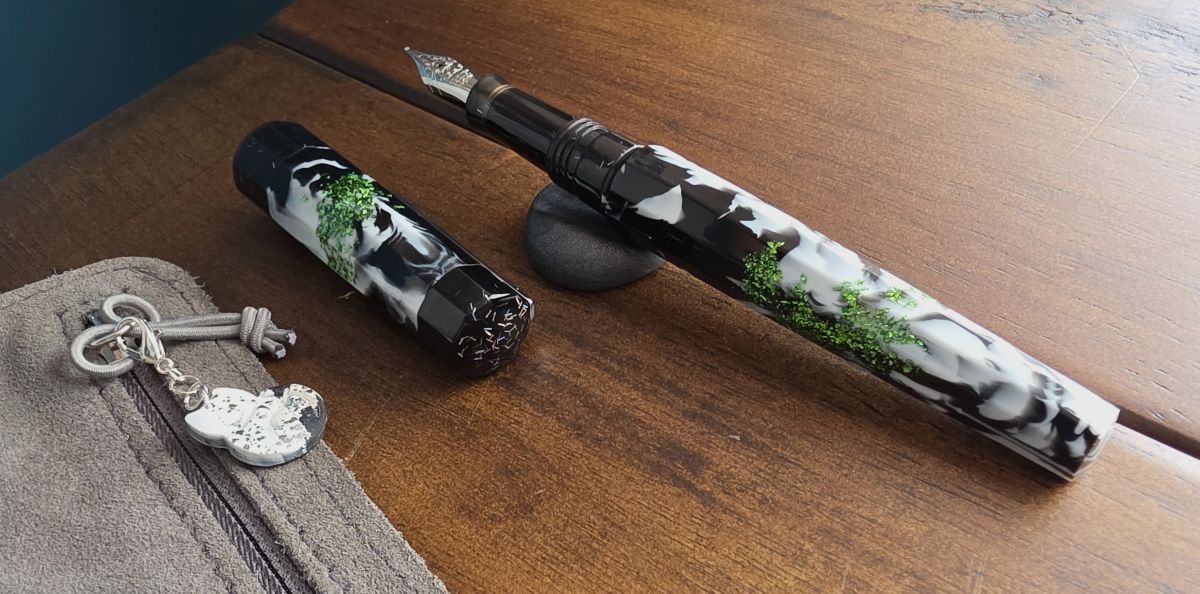 An uncapped Benu Euphoria pen with black and white swirled resin, patches of shiny bright green and holographic bar glitter in the cap and section. A black and white cat charm with silver glitter clipped to the pull of a zipper pocket.