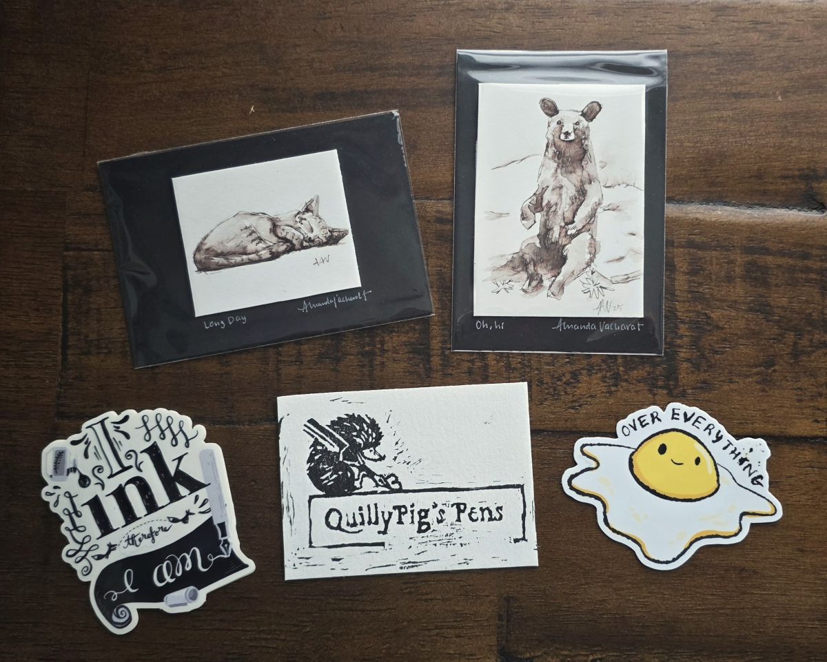 Ink painting prints of a sleeping cat and a sitting bear, a sticker with text that says I ink therefore I am, a sticker of an egg with the text Over Everything, and a cart with a carved design of a hedgehog with a pen for QuillyPig's Pens.