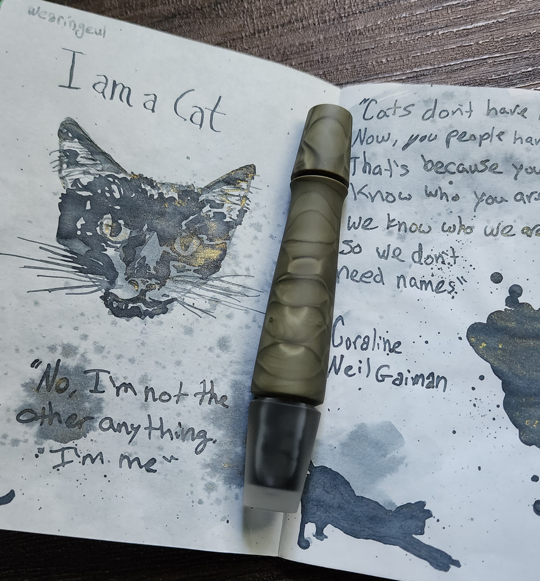A short, curvy fountain pen with a small stopper-style cap in army green swirl and transparent resin with a matte finish, resting on an open notebook with the I am a Cat ink and sketch of Boo.