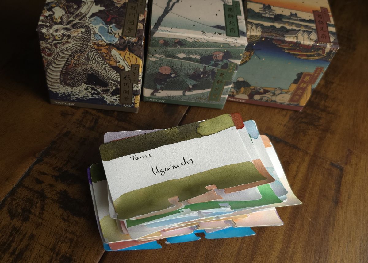 Stack of Col-o-dex cards with ink swatches, a dark olive green called Taccia Uguisucha is on top with three ornate Taccia ink boxes in the background.