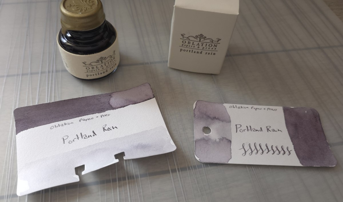 A bottle of Oblation Paper and Press Portland Rain Fountain Pen ink next to Col-o-dex and Col-o-ring cards swatched with the ink which is a dusty gray purple shade.