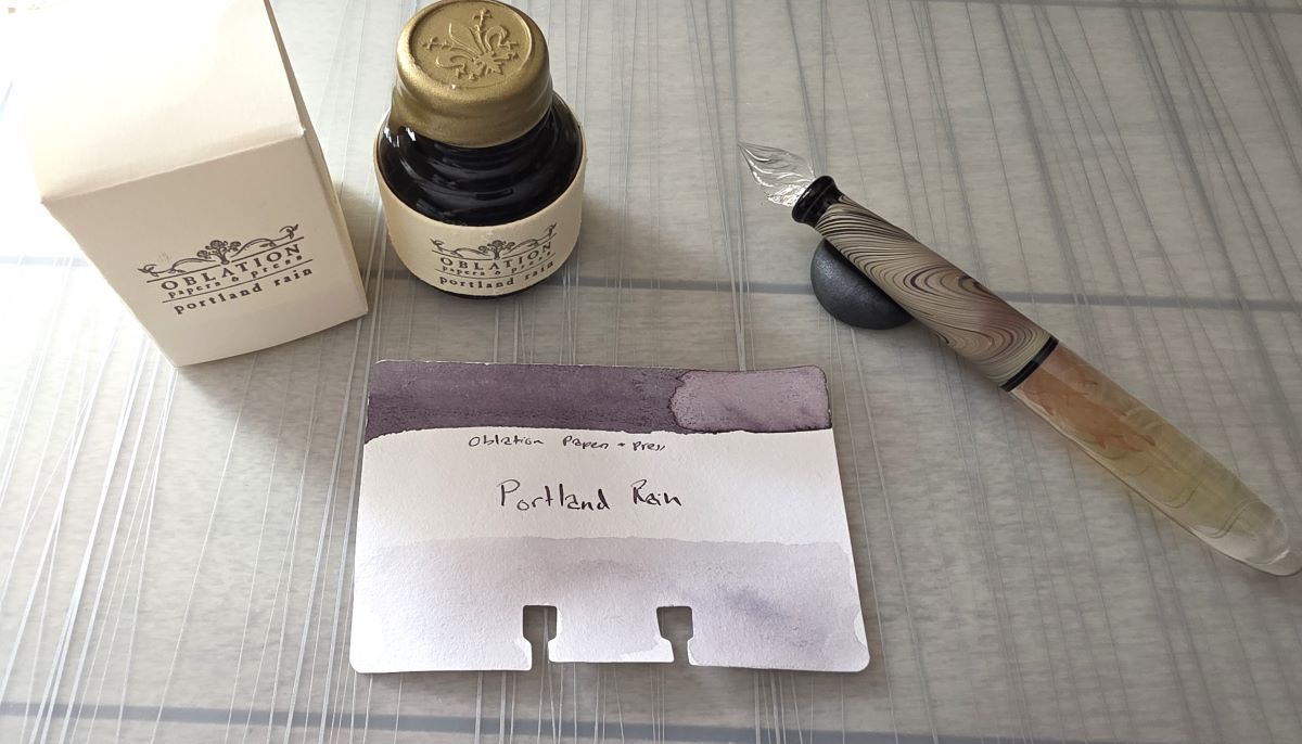 A bottle of Oblation Paper and Press Portland Rain Fountain Pen ink next to a Col-o-dex card swatched with the ink which is a dusty gray purple shade and an ornate handmade glass pen resting in a pen stand.