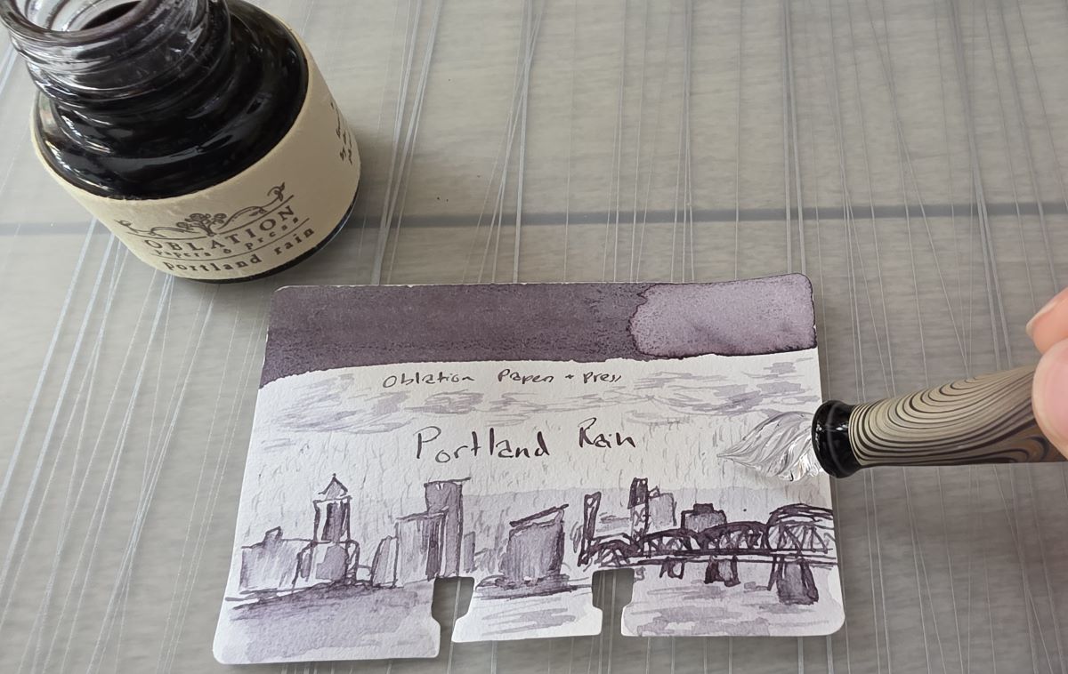 A glass pen with diluted Portland Rain ink being used to sketch in dark clouds and rain falling onto the skyline.
