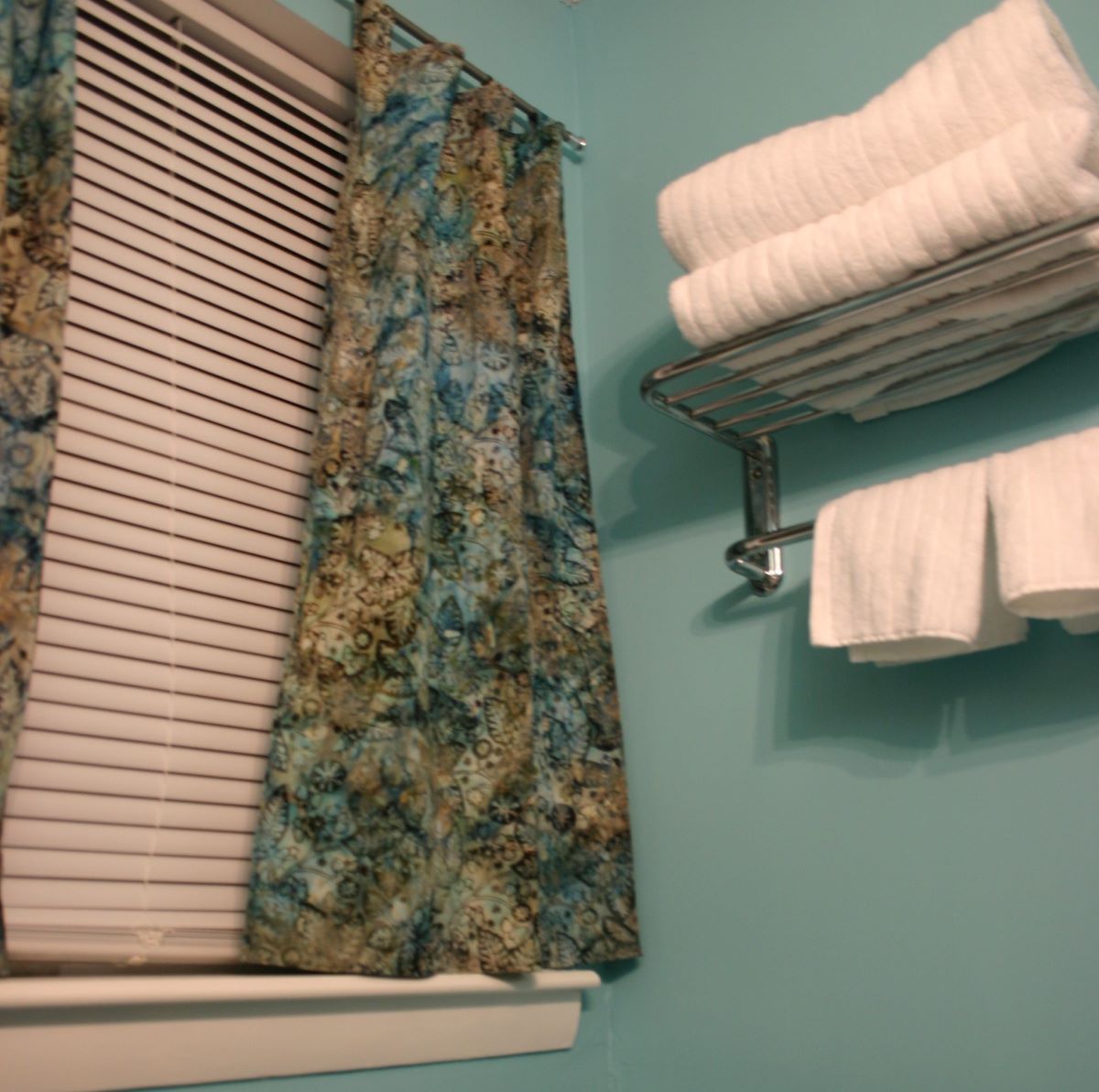 Corner of bathroom wall with light blue paint, blue and brown batik curtains, and fluffy white towels on a rack on the wall.