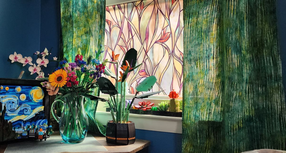 A bright window with window film that looks like stained glass, green and blue batik curtains, medium blue wall, and a small desk with several Lego sets including an orchid, wildflower bouquet, Van Gogh painting, bird of paradise, and succulents.