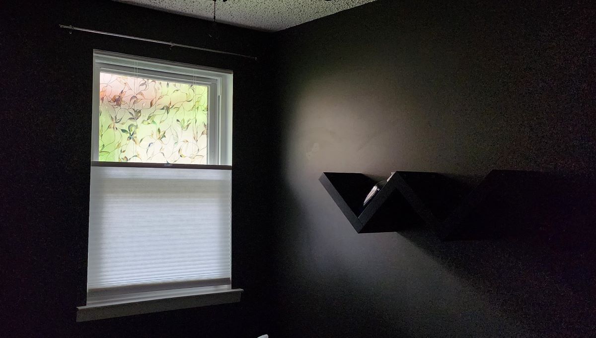 The corner of a room painted dark gray, a window with a frosted glass look window film on the top half and a top-down fabric blind covering the lower half of the window.