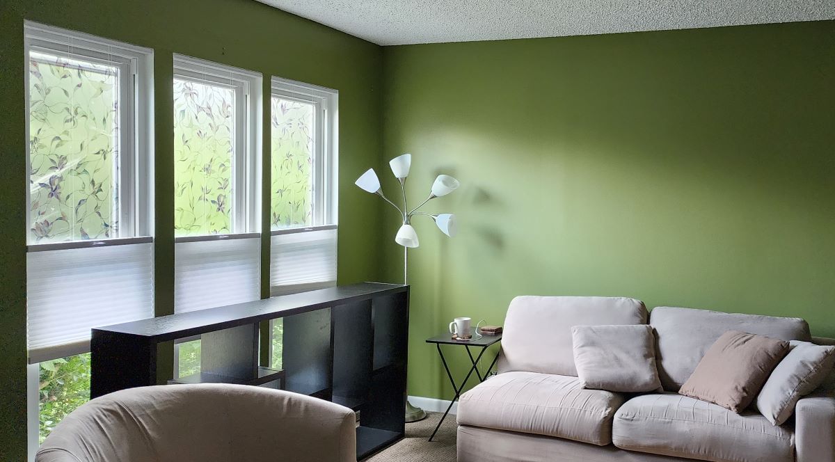 A room painted a medium dusty green with bright narrow windows, a neutral color couch and carpet, and floor lamp with five bulbs and shades.