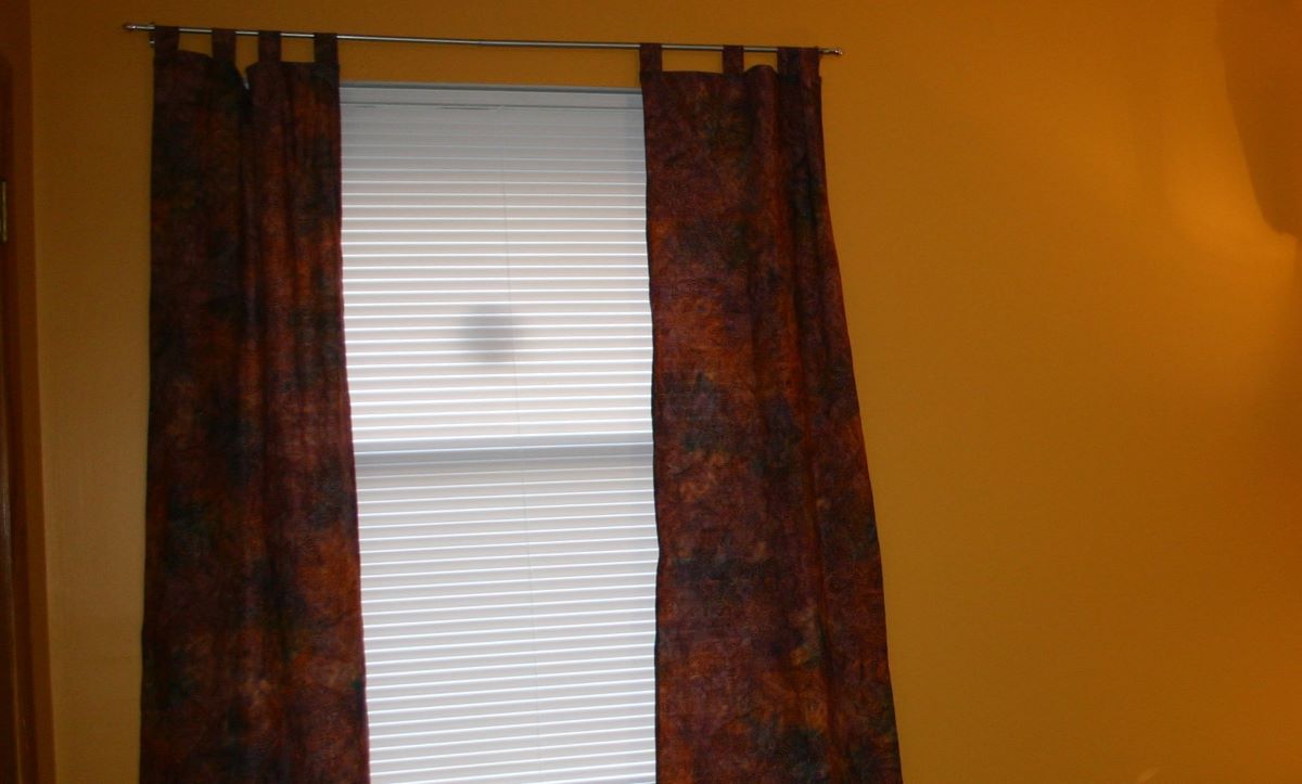 A wall painted orange, window with a white blind and purple and orange batik curtains along the sides.