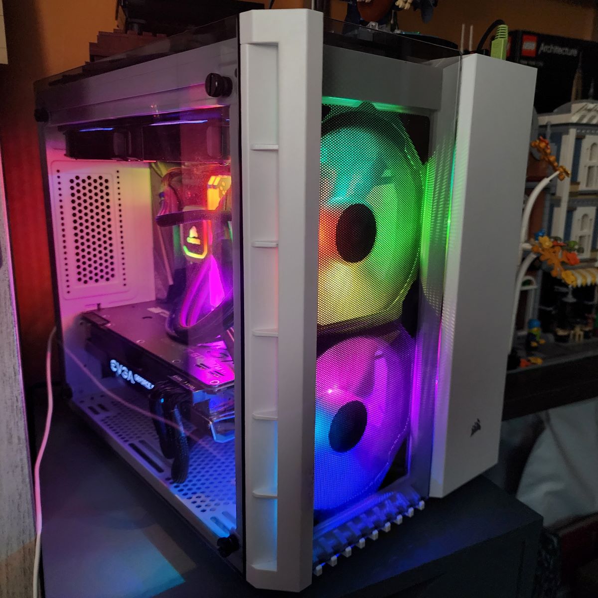 A slightly square PC computer case in white with large glass panels on the front, side, and top filled with rainbow-colored glowing RGB lights on the fans, memory stick, cooler, etc.