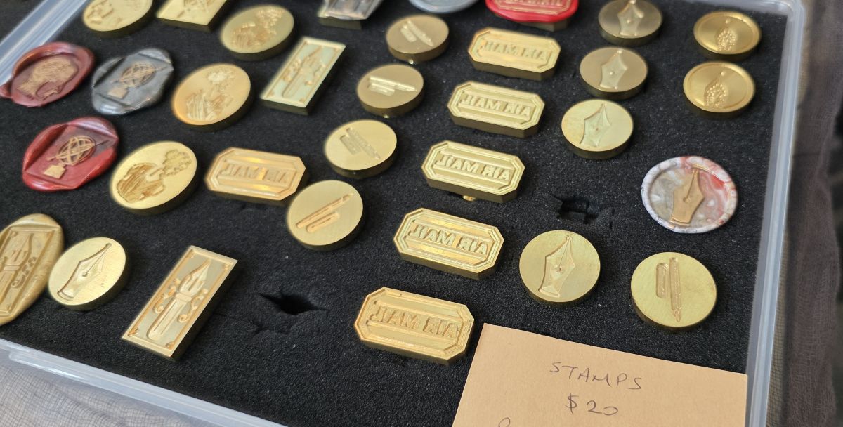 Closeup of a display on a table at a pen show of several metal wax seals with fountain pen nib, fountain pen, and air mail theme designs.