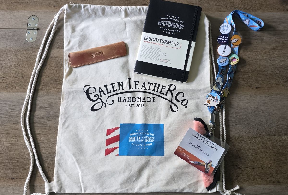 A large natural canvas drawstring backpack with Galen Leather Co and Washington DC Supershow logos printed on the side, a light brown Galen leather single pen sleeve, an A5 Leuchtturm1917 black notebook with the pen show logo on the cover, a lanyard covered with pins and buttons with a fuzzy pink Rickshaw single pen sleeve and show badge for Lisa, OliveOctopus.Ink on the clip.