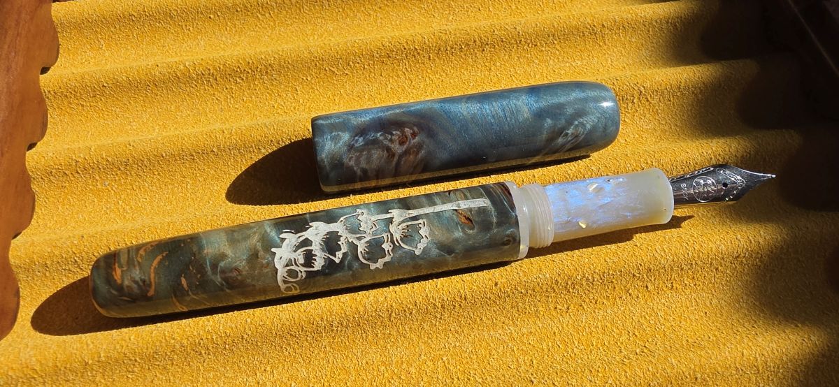 An uncapped fountain pen made of wood and resin in dusty shades of blue, green, and orange with a carving in the body of a lily of the valley flower painted white, a sparkly pearlescent white section, and medium Bock nib, resting in a yellow pen tray in bright sunlight.