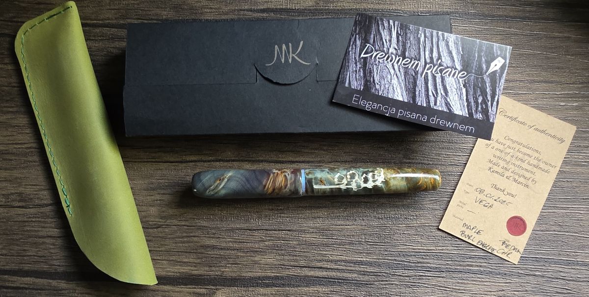 A fountain pen made of wood and resin in dusty shades of blue, green, and orange with a carving of a lily of the valley flower in the body painted white, a spring green leather single pen sleeve, black paper pen box, business card for Drewnem pisane, and card with details about the pen model and materials.