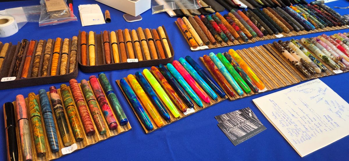 A table at a pen show with trays holding dozens of handmade fountain pens made of natural and dyed woods, oparex, resin with dried flowers or coffee beans, other resins, etc.