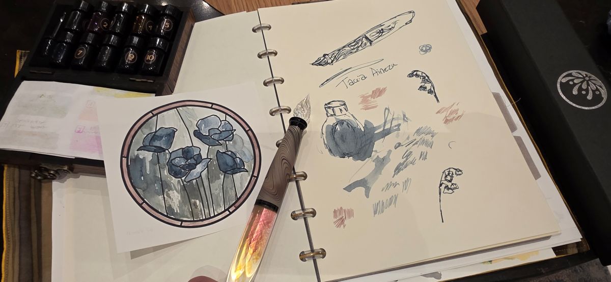 A glass dip pen from Hanabi with swirled wood-look grip and colorful twists in the body resting on a disc-bound notebook with random writing and sketches, an image of stained glass flowers colored in blue, green, and light brown, and a small wooden case with 10 tiny glass bottles of fountain pen ink.