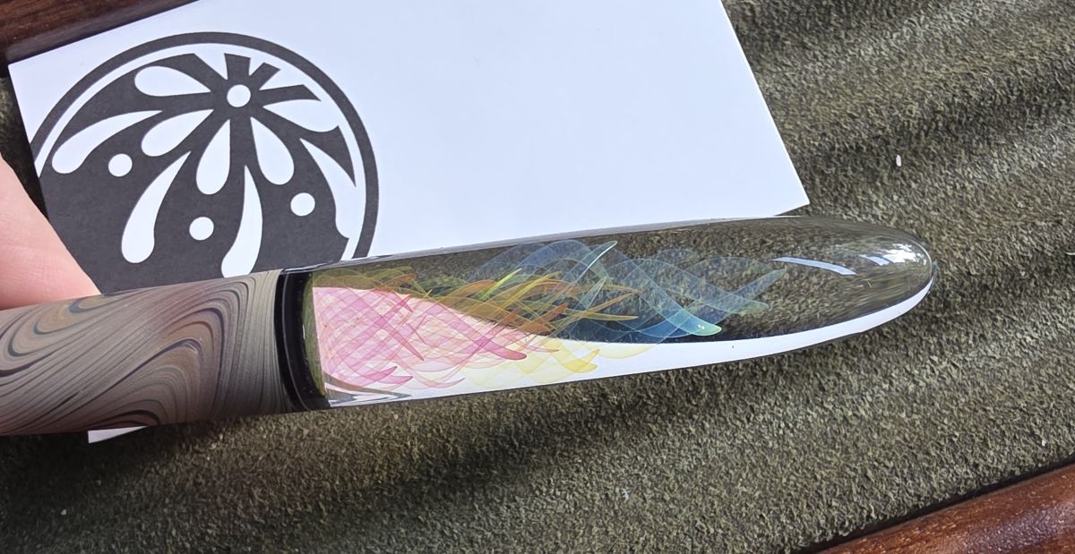 The handle of a glass dip pen with part of the glass reflecting a white background and the colorful swirls looking pink and yellow and the rest reflecting dark green background where the same swirls look yellow/orange and blue/green respectively.