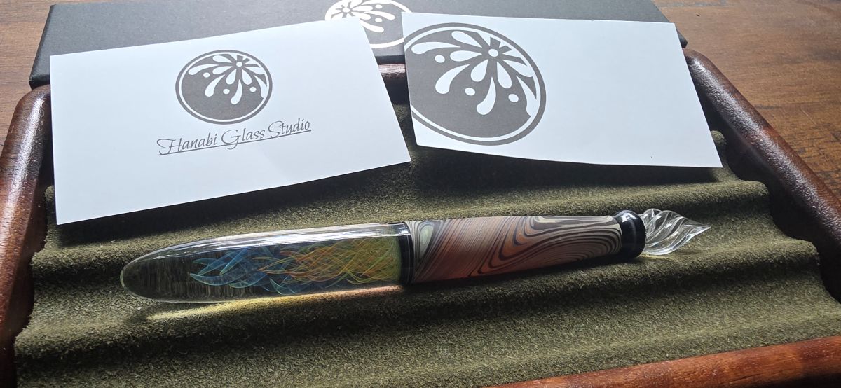 A glass dip pen with twisted channels and a fine tip, a matte finish grip area that looks a bit like wood, and colorful swirls of blue and orange in the handle, resting in a dark green pen tray next to business cards with the Hanabi logo.