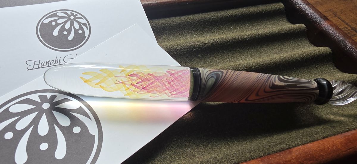 The handle of a glass dip pen over white cards with the Hanabi logo that make the colorful swirls look pink and yellow.