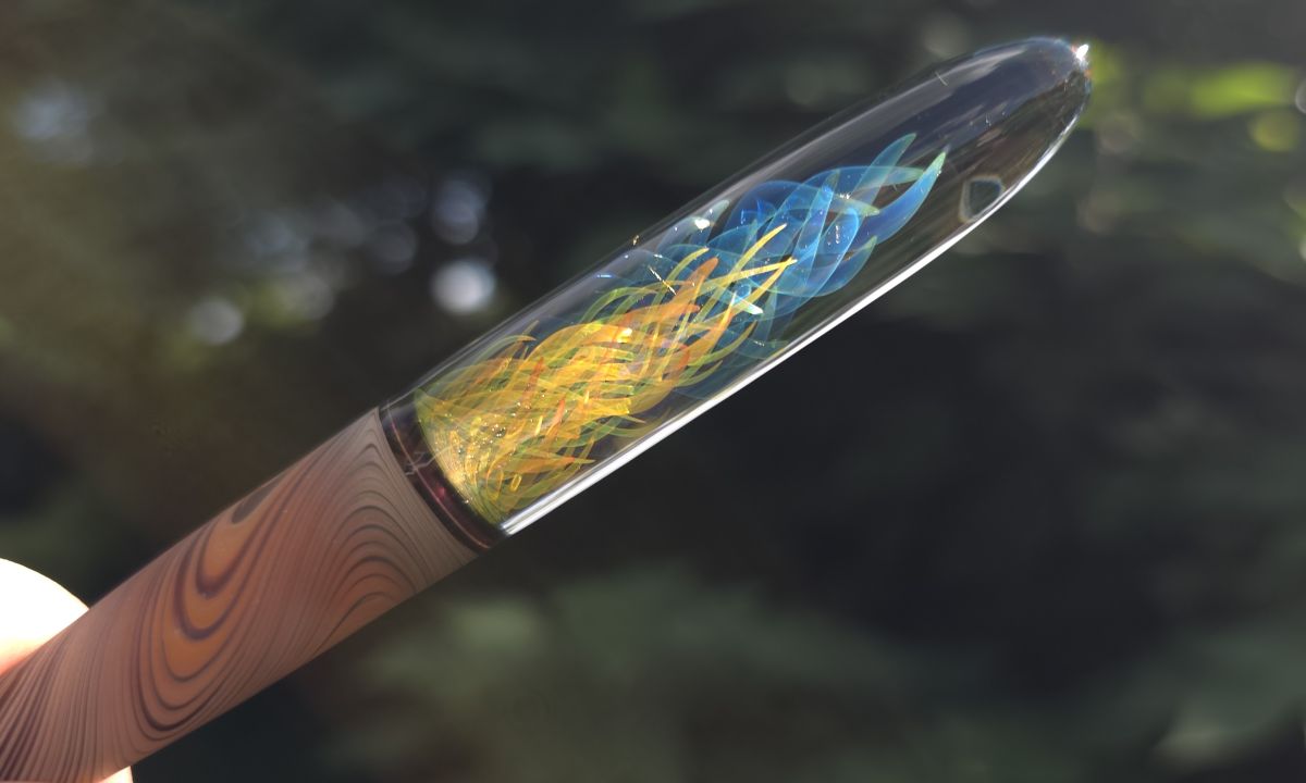 The handle of a glass dip pen held up in the sunlight with dark foliage out of focus in the background and large swirls of blue and green and finer swirls of yellow and orange glistening in the sunlight.