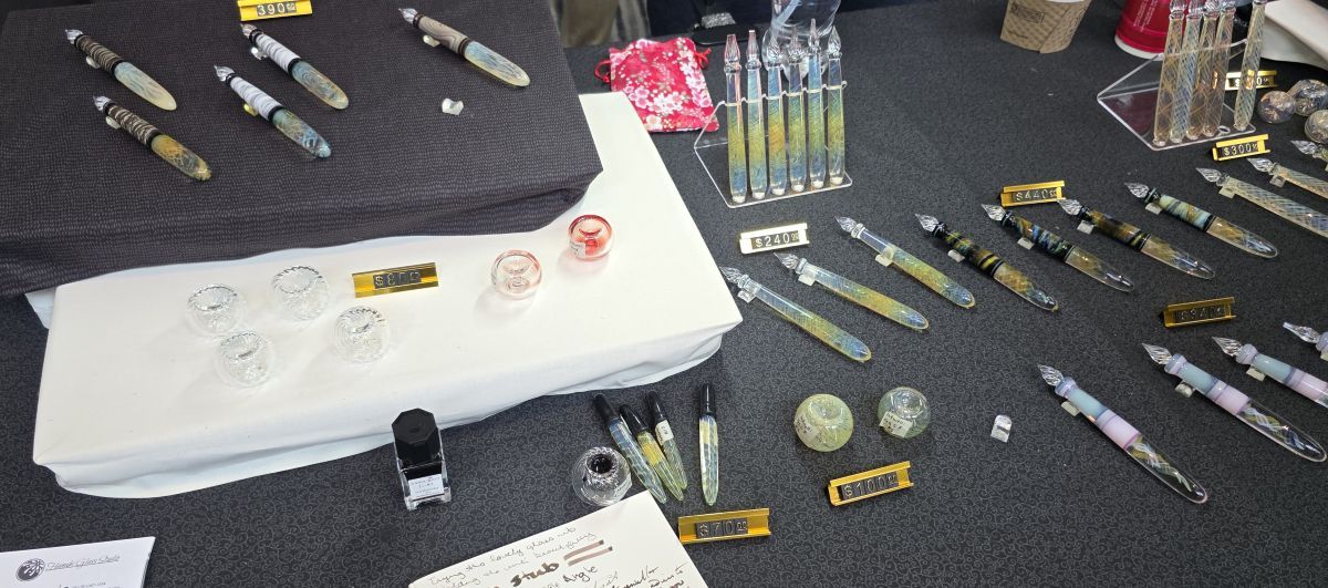 A table at a pen show for Hanabi Glass with several colorful glass dip pens, eye droppers, and ink wells.