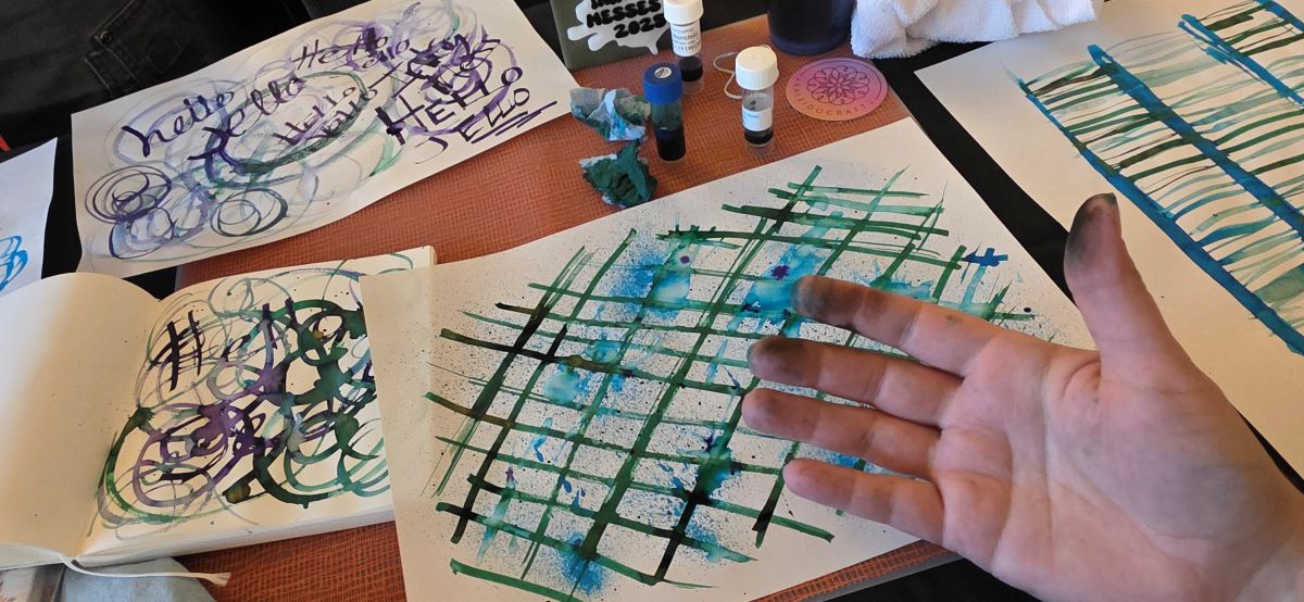 A hand with slightly inky fingers over several sheets of paper with colorful ink lines, mist, splats, circles, words, etc. in blue, green, and purple.