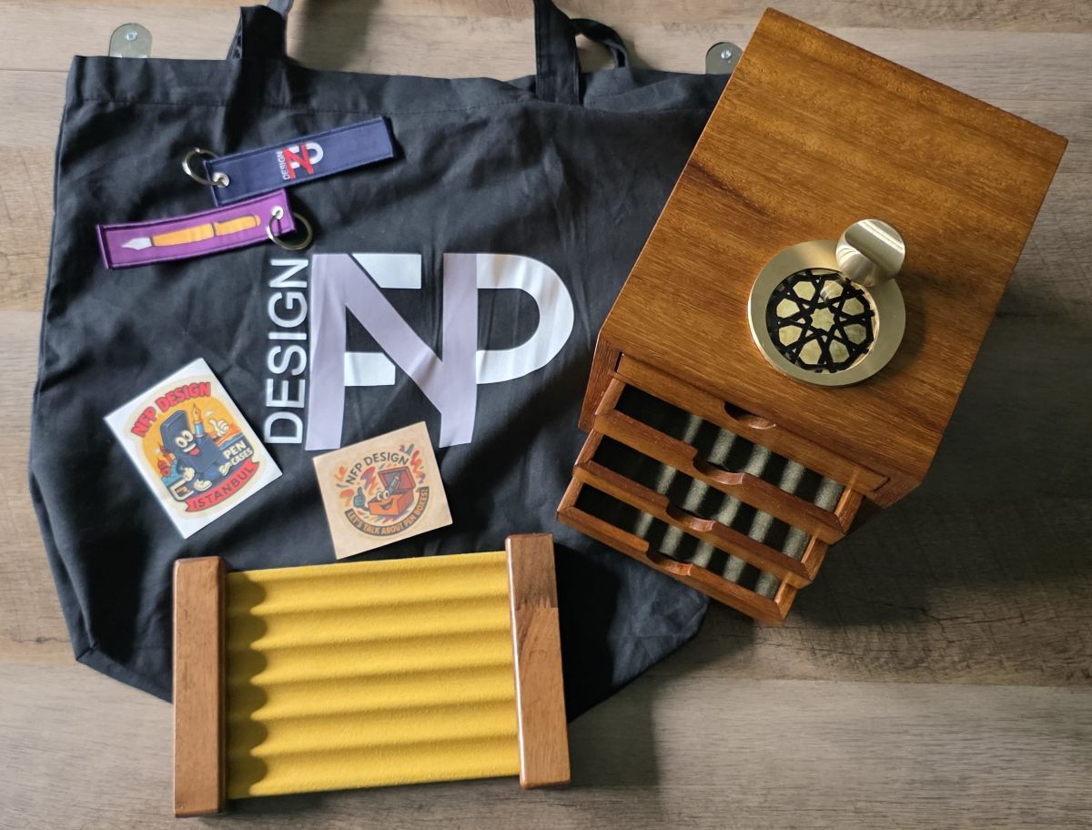 A large black cloth bag with NFP Design logo, two keychains with NFP logo and fountain pen graphic, NFP stickers, a yellow 6 pen tray suspended between wood supports on the ends, a compact wooden pen storage box with four drawers that each hold 6 pens with a dark green liner, a small brass pen rest with a dark yellow and black pattern tile decoration.