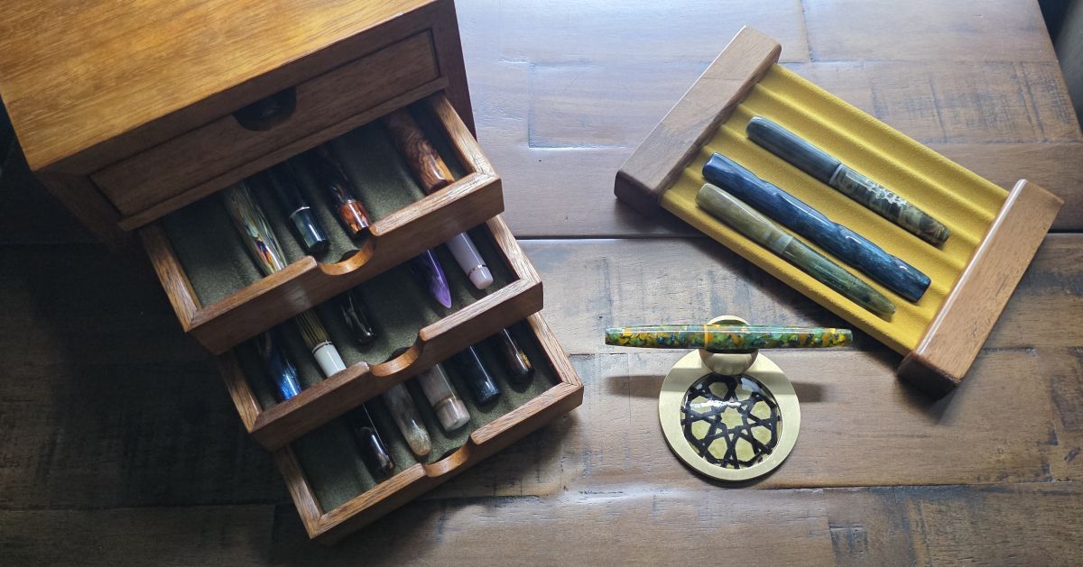 Three pen storage items from NFP holding a variety of fountain pens including a compact wooden pen storage box with four drawers with dark green pen tray liners that can each hold 6 pens, a small brass pen rest with a dark yellow and black pattern round tile decoration and pen held in a slot in a ball of brass attached to the edge, a yellow 6 pen tray suspended between wood supports on the ends.