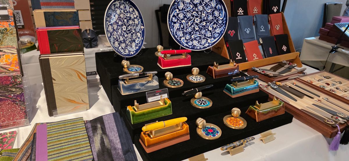 A table at a pen show with fabric covered notebooks, large patterned ceramic plates, two styles of ceramic and brass single pen rests, pen cases and sleeves, and other tile items for sale.