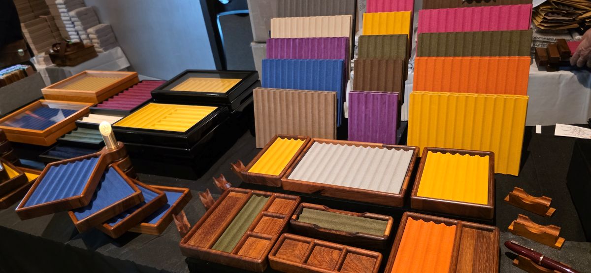 A table at a pen show with pen storage trays made of wood in various shapes and sizes with slotted pen tray inserts in shades of dark green, orange, blue, yellow, tan, cream, gray, brown, pink, and purple.
