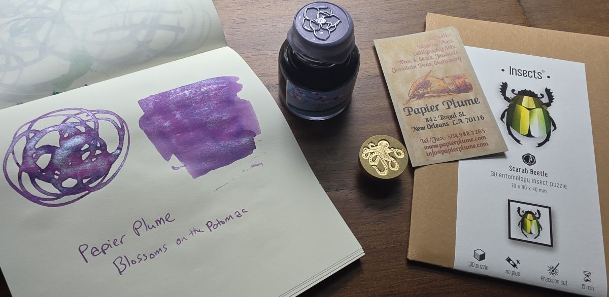 A bottle of fountain pen ink with a light purple wax seal and flower on the cap, a notebook with a swatch of the ink which is a medium chromashading purple with lilac shimmer, an envelope with a paper scarab bug puzzle, and a metal wax seal with an octopus design.