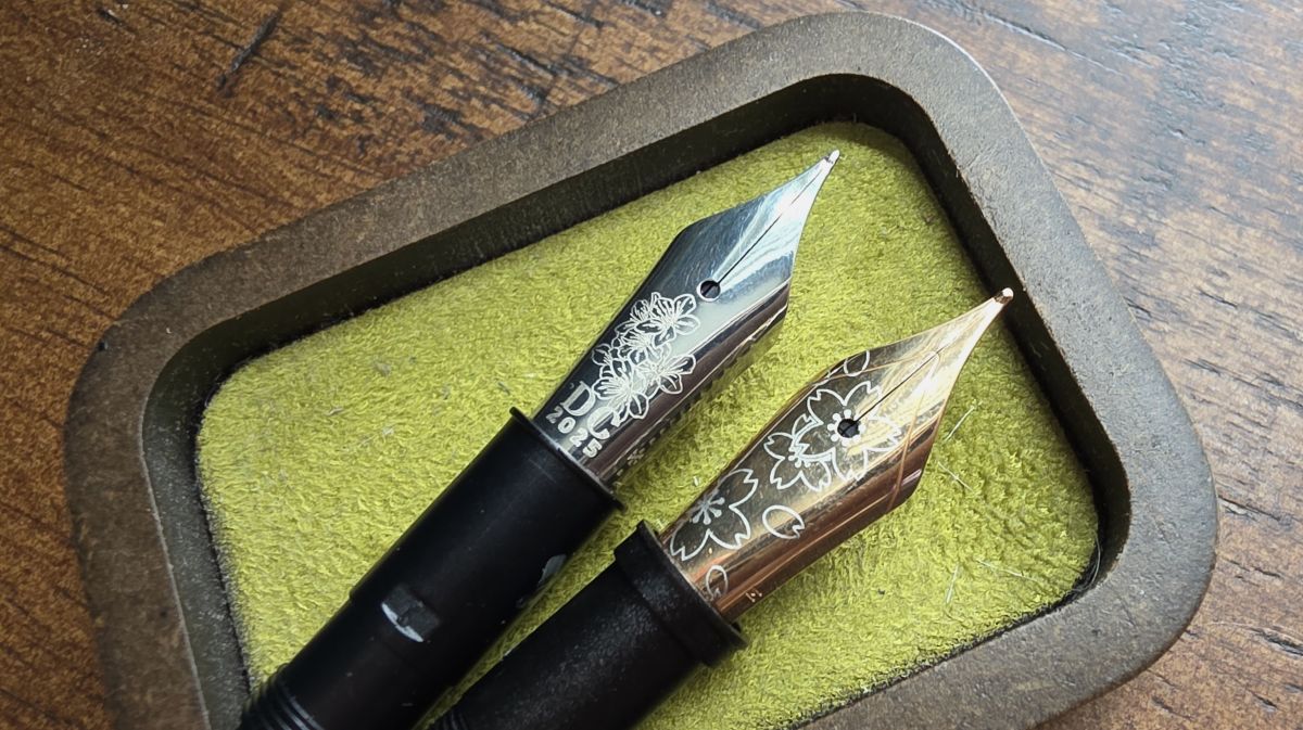A close up of two loose fountain pen nibs in a small wooden, fabric-lined tray, one medium Bock nib with a cherry blossom design and a medium Jowo nib in rose gold color with sakura blossoms.