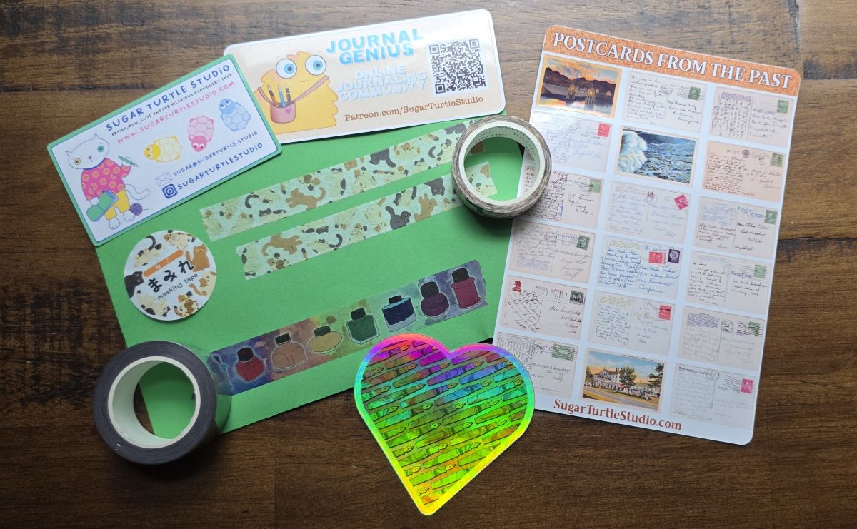 A sheet of stickers with photos of old used postcards, a large iridescent heart sticker with fountain pen design, Two rolls of washi tape and a large green envelope with swatches of washi tape with cats and a rainbow of fountain pen ink bottles, and a couple cards for Sugar Turtle Studio and the Journal Genius Patreon community.