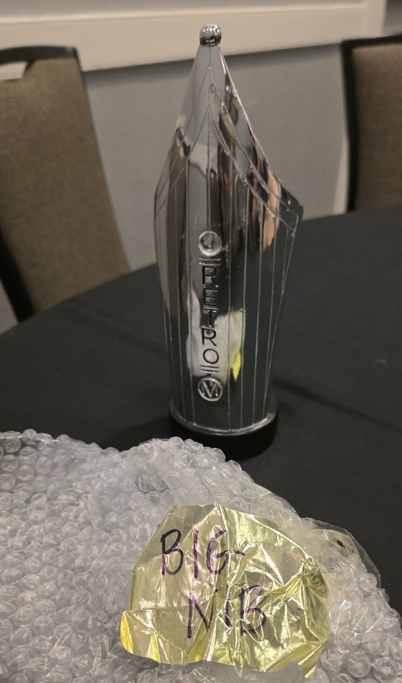 An oversized model of a Retro 51 fountain pen nib, nearly a foot tall, used to identify the main parts of a nib, next to packing material with a label that says 'Big Nib'.