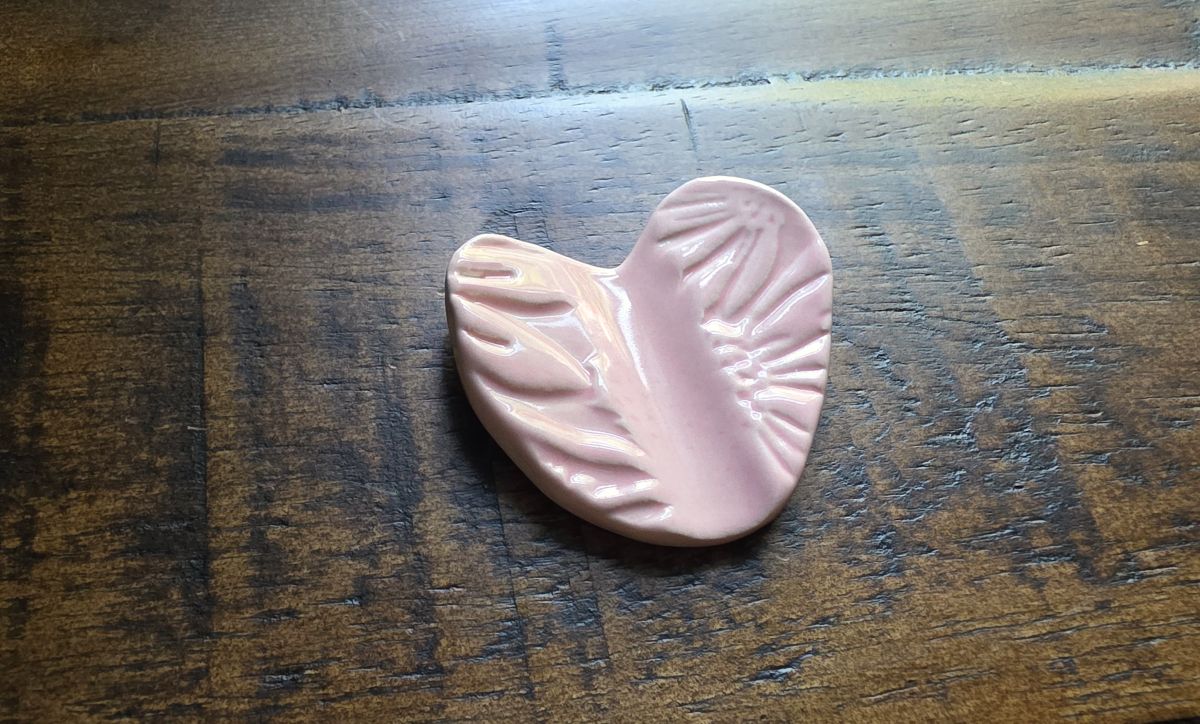 A small ceramic pen rest in the shape of a heart with floral design pressed into the top in a light pink color.