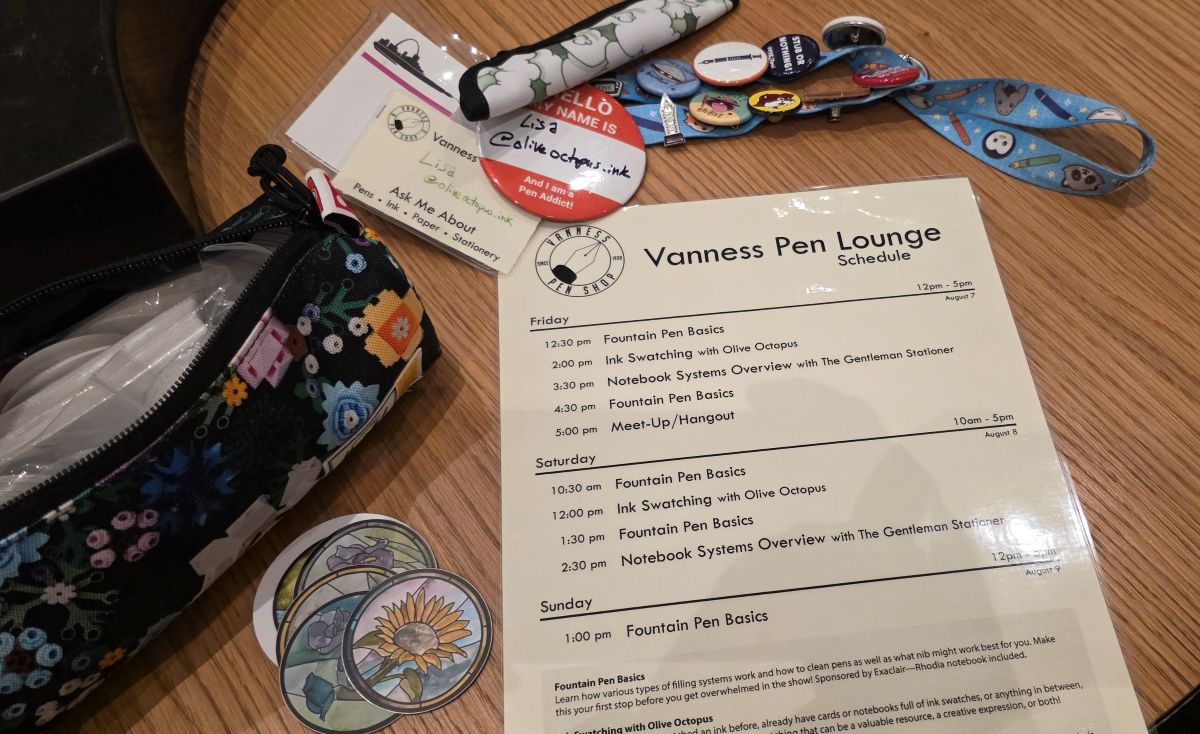 A table covered in a pouch with stickers, a lanyard covered in pins and buttons with name tag for the pen show, and laminated sheet with the Vanness Pen Lounge schedule.