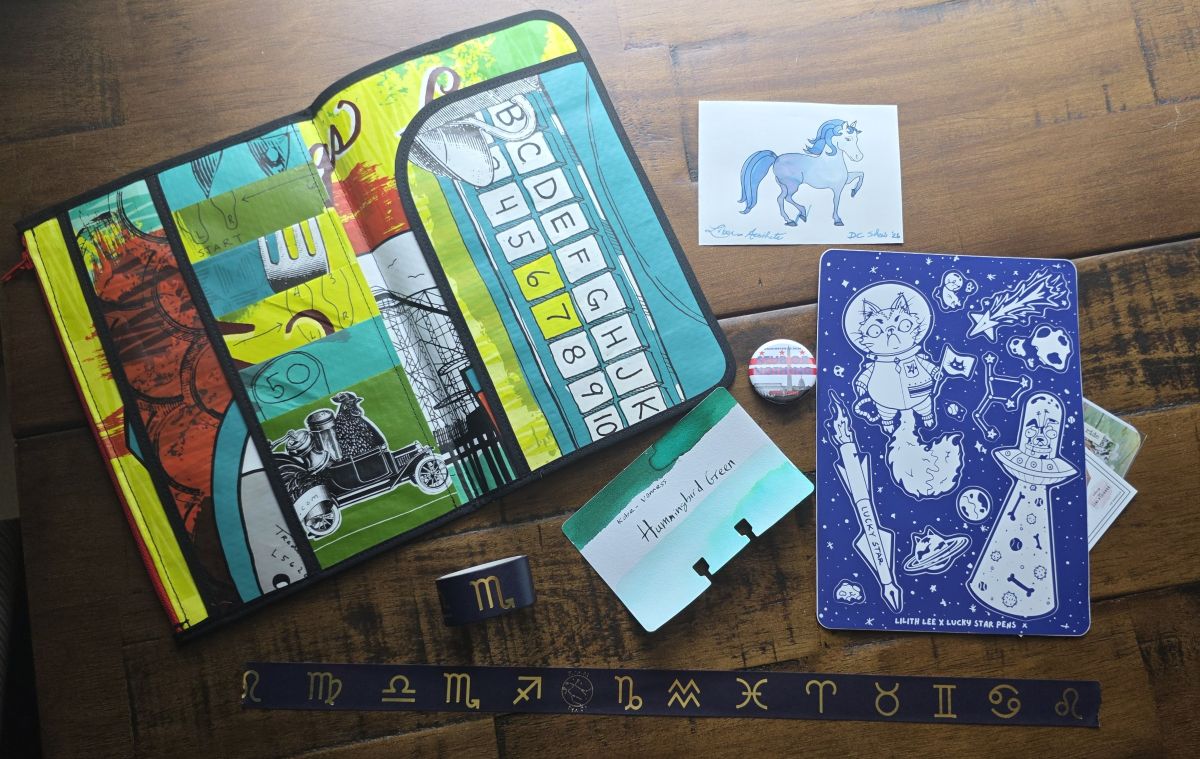 An A5 zipper pouch/notebook folder made from a reusable shopping bag, a painted ink pony, stickers and trading cards, button, ink swatch card for Kobe Vanness Hummingbird Green, and a roll of zodiac washi tape.