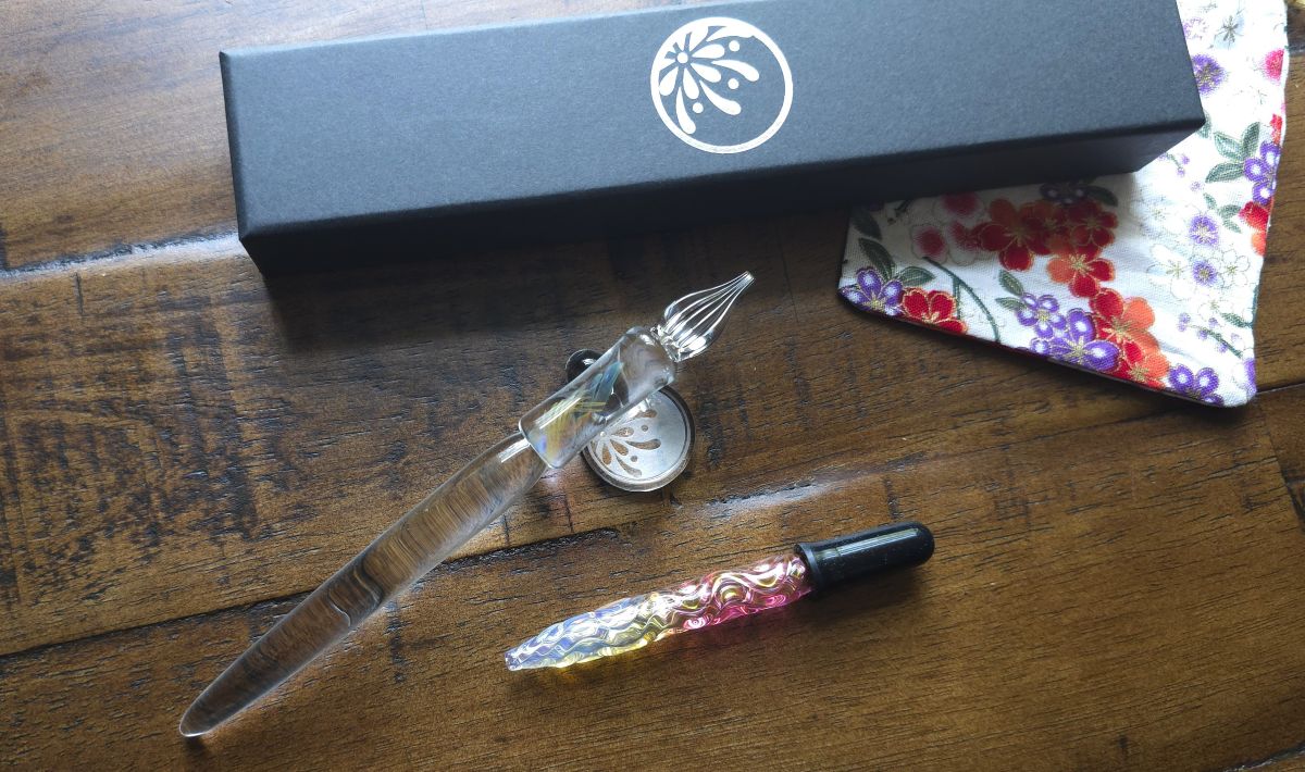 A hand-made glass pen with a clear handle, shaped grip section and colorful swirl, and an italic style tip, and a colorful glass eyedropper, both from Hanabi Glass Studio.