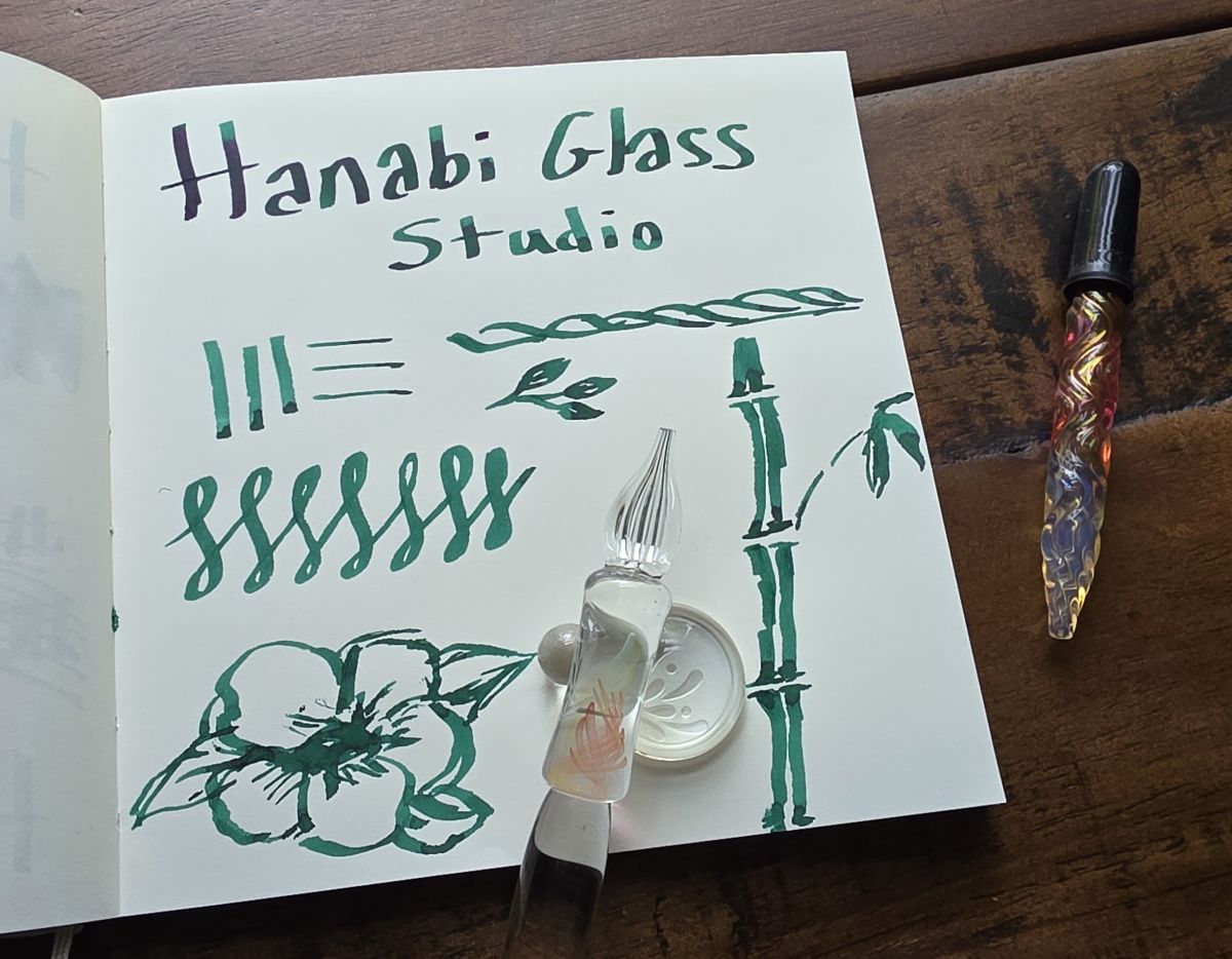 A page of writing and sketching with an italic-style glass pen, the glass pen on a glass pen rest with the Hanabi logo, and a colorful glass eyedropper.