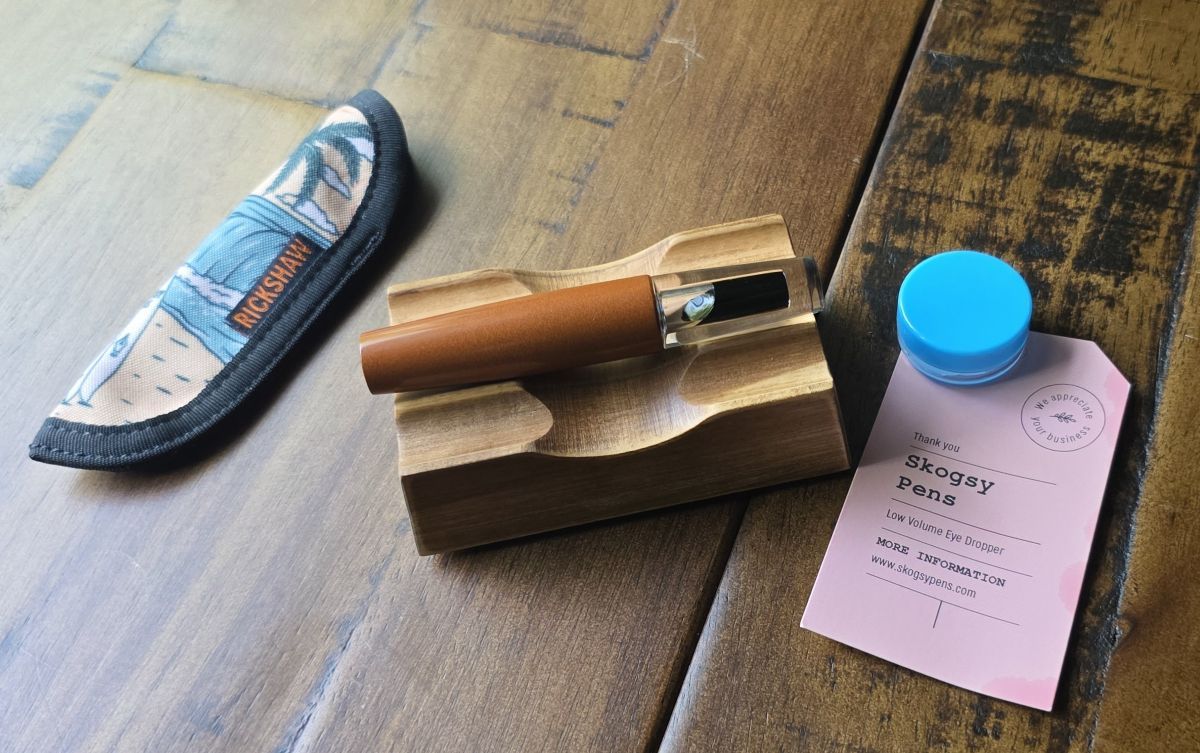 A pocket-size Skogsy LVED fountain pen with orange brown ebonite cap filled with dark orange ink, next to a small Rickshaw pen sleeve, silicone grease, and information card.