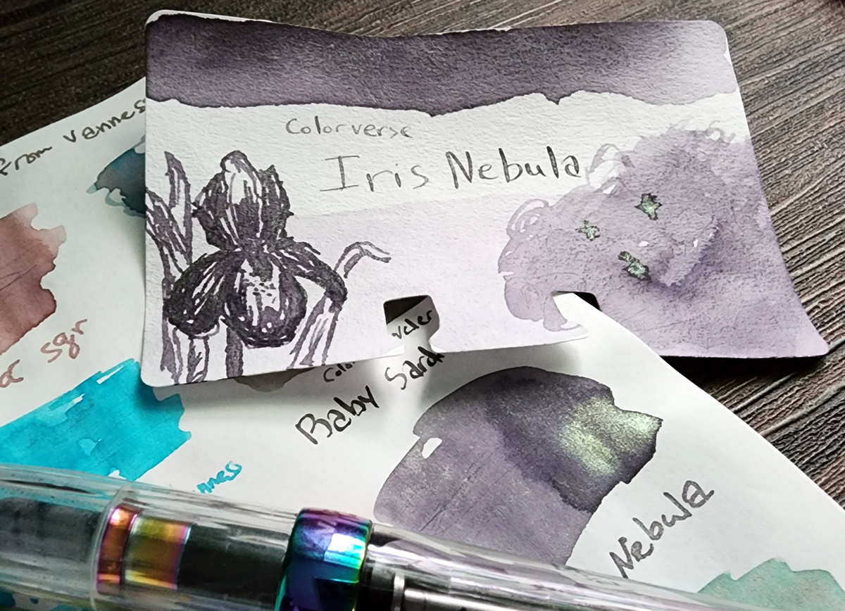 A Col-o-dex swatch card and swatch on Tomoe River paper for Colorverse Iris Nebula, a dusky purple with gold/green/blue shimmer, next to the rainbow anodized TWSBI Vac 700R Iris pen.