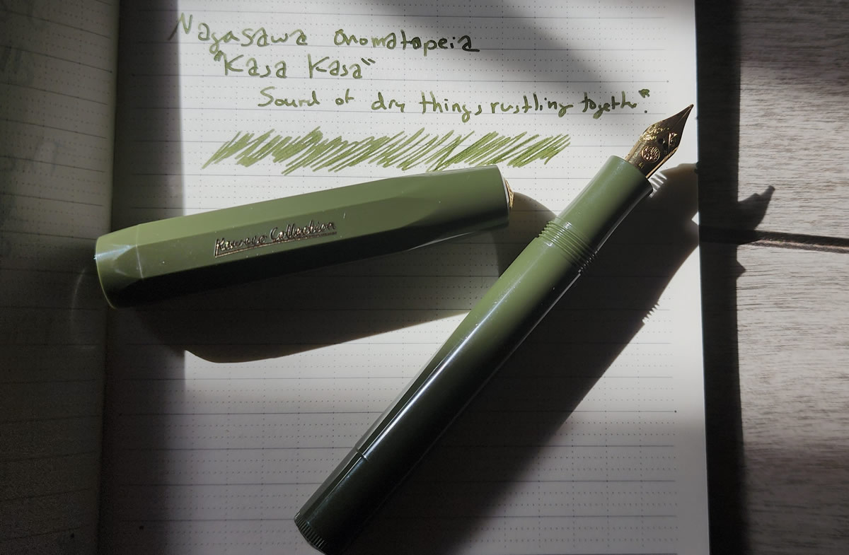 A Kaweco Sport pen in dark olive on a notebook with a writing sample of the matching green Nagasawa Kasa Kasa ink illuminated in a sunbeam hitting the desk.