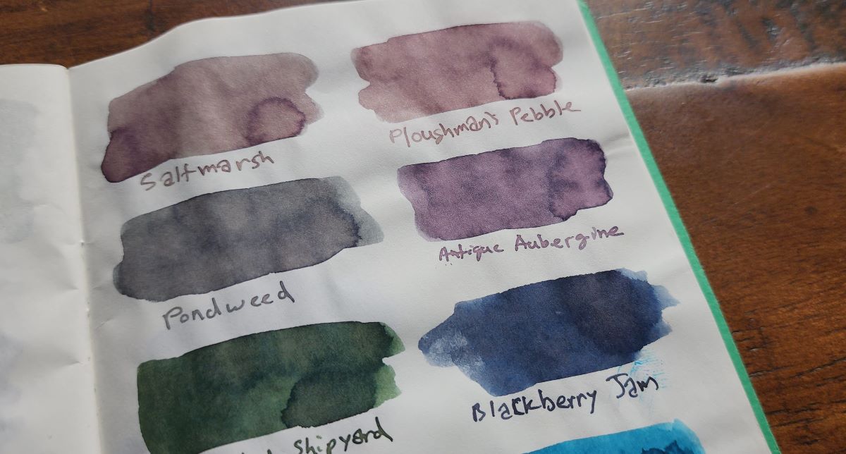 A notebook page with six ink swatches for Birmingham Pen Company inks Saltmarsh, Ploughman's Pebble, Pondweed, Antique Aubergine, Abandoned Shipyard, and Blackberry Jam. Antique Aubergine is a dusty gray pink purple shade.