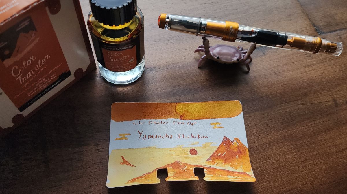 Glass ink bottle, box, and Col-o-dex ink swatch card with sketch of sunset and mountains for Color Traveler Yamanohaitochikou, a rich shading yellow orange. TWSBI Diamond 580 Sunset Yellow pen filled with that ink, held by a purple crab pen holder.