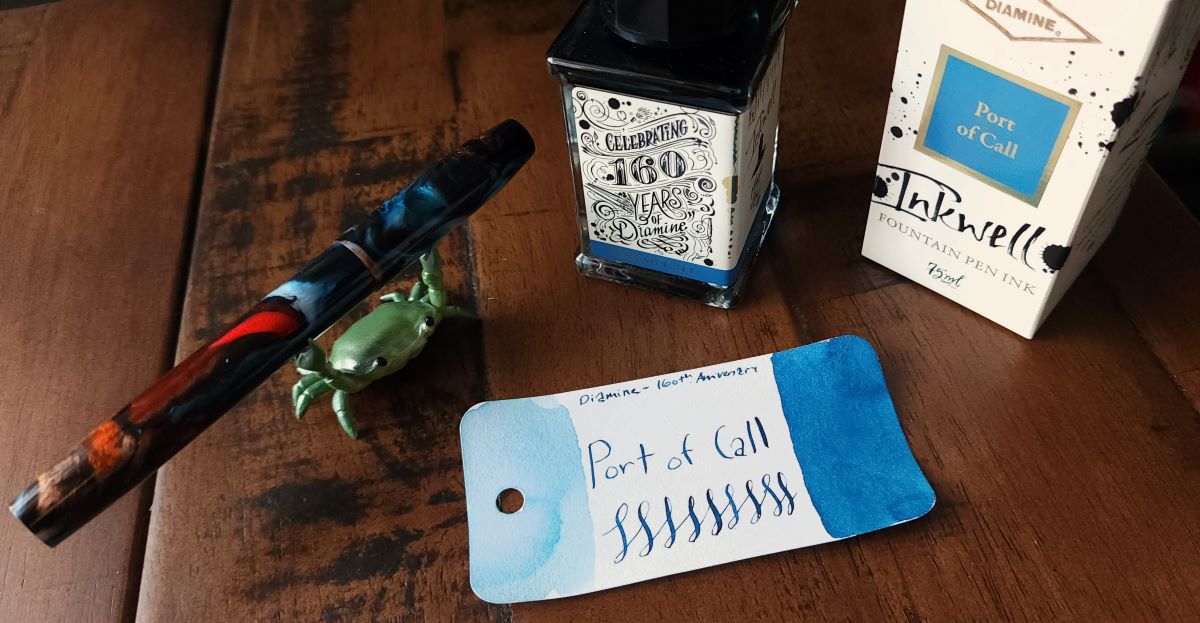 A tall rectangular glass ink bottle and Col-o-ring swatch card for Diamine 160th Anniversary Port of Call, a medium blue. Franklin Christoph Model 46 fountain pen made with swirly transparent blue, transparent orange, black, and white resin, held by a green crab pen holder.