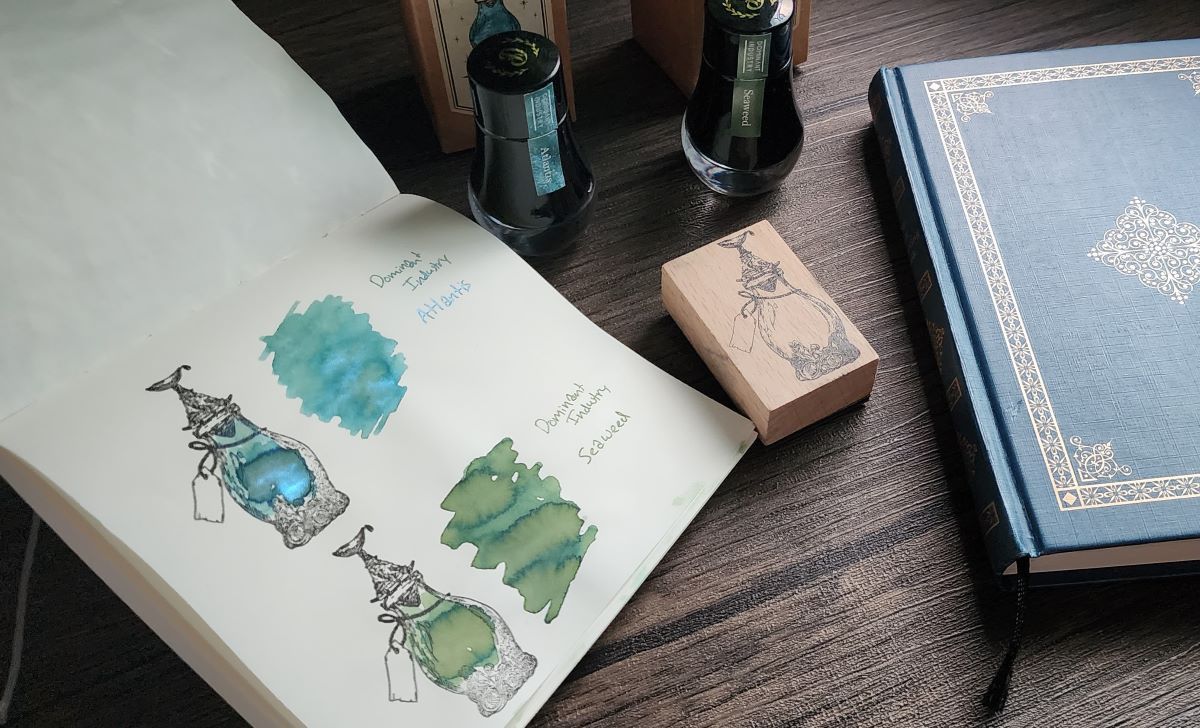 Two bottles and swatches of Dominant Industry fountain pen ink, a blue-green with bright blue shimmer called Atlantis, and a chromashading green called Seaweed. A rubber stamp of an ornate bottle. The Dominant Industry Ink Archiving Book, a B6 hardcover with dark blue color and foil design. 