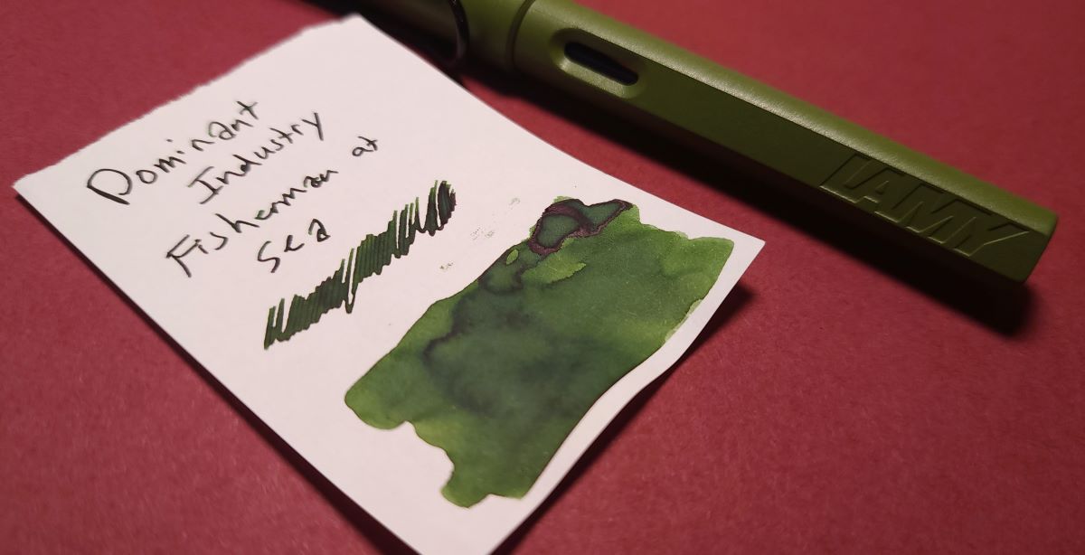 Ink swatch for Dominant Industry Fisherman at Sea, a dark yellow green with slight red sheen, next to a Lamy Safari in Savannah Green.