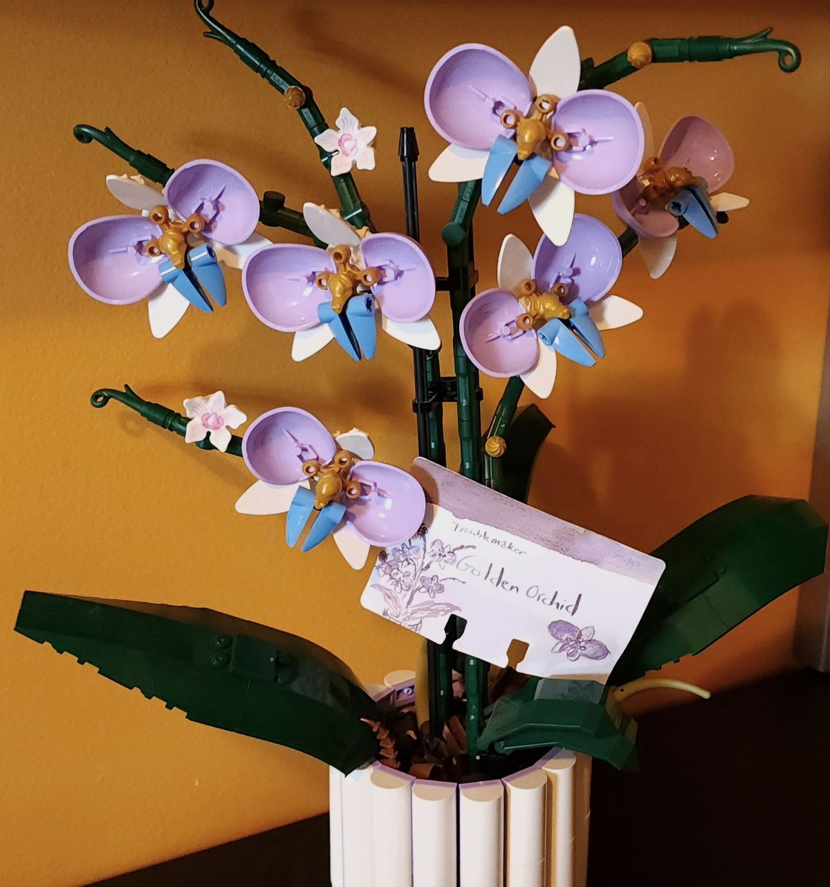 A Lego Orchid set that has been customized with lavender, gold, and periwinkle flowers and a white pot and the Col-o-dex ink swatch card for Troublemaker Golden Orchid tucked in between a flower and dark green leaves.