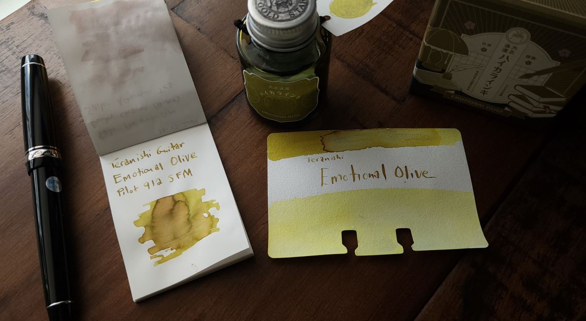 Bottle of Teranishi Emotional Olive fountain pen ink, Col-o-dex swatch card showing the yellow and brownish green shade, small notebook with swatch and writing sample from the black Pilot Custom Heritage 912 pen with a Soft Fine Medium nib next to the notebook.