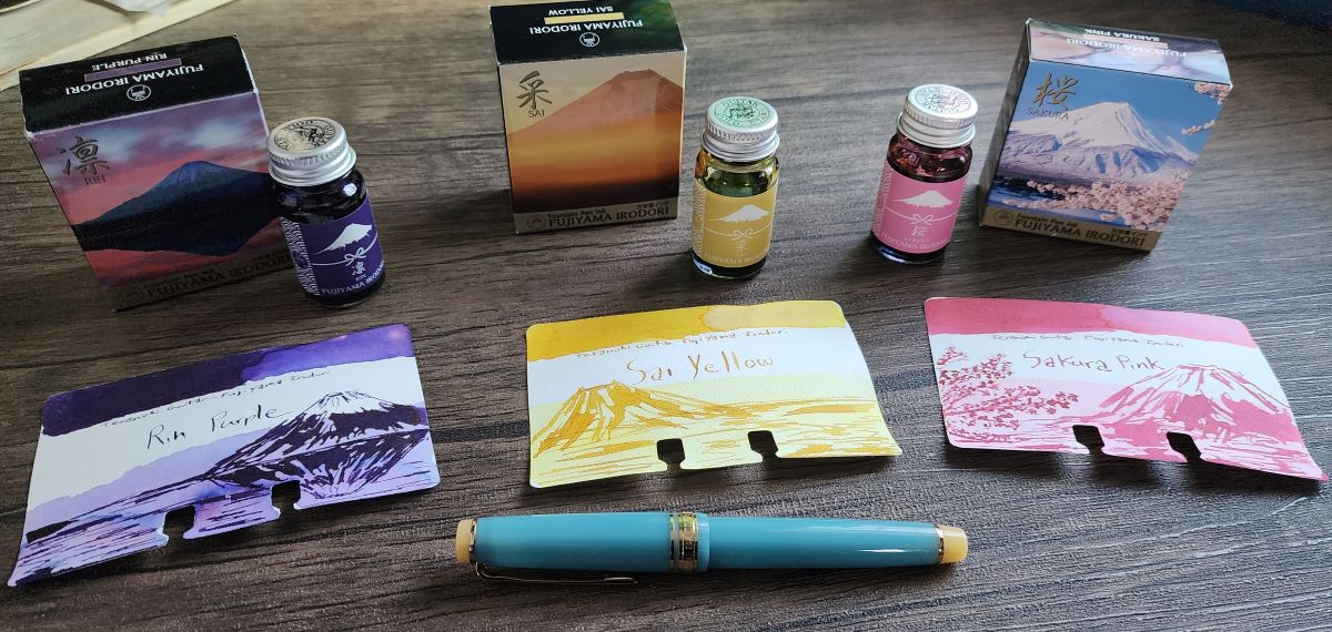 Three small bottles of Teranishi fountain pen ink inspired by views of Mount Fuji: Rin Purple for the dark purple mountain and a pink sky, Sai Yellow for a warm yellow sunset, and Sakura Pink for pink sakura blossoms in front of the snowy peak. Three corresponding Col-o-dex swatch cards with sketches of Mount Fuji.