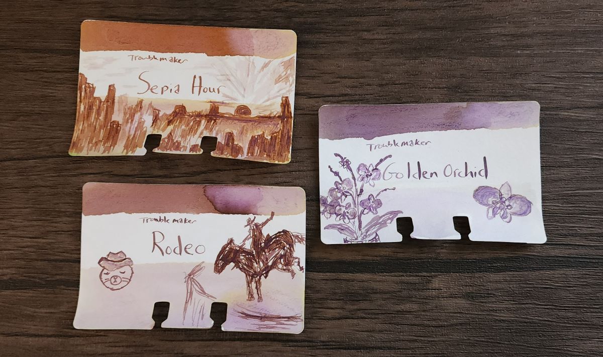 Three Col-o-dex ink swatch cards for Troublemaker inks Sepia Hour, a rusty orange with purple shading, Rodeo, a mix of dusty purple and rusty orange, and Golden Orchid, a dusty purple with slight golden shading.